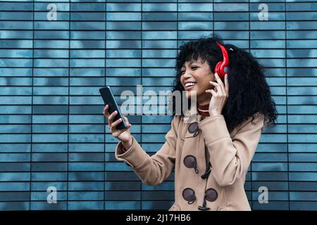 Cheerful young woman wearing wireless headphones using smart phone in front of blue wall Stock Photo