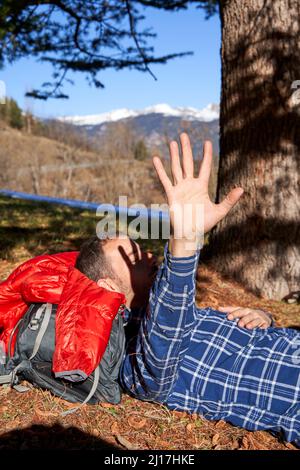 Man raising his hand lying under tree on sunny day Stock Photo