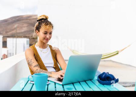 Smiling young woman using laptop sitting at table on rooftop Stock Photo