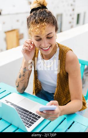 Happy young woman surfing net through mobile phone sitting at table on rooftop Stock Photo