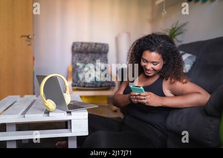 Smiling woman text messaging through smart phone sitting by sofa at home Stock Photo