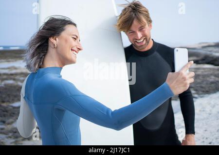 Happy couple in wetsuit  taking selfie with surfboard at beach Stock Photo