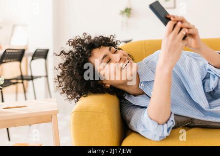Happy young woman text messaging through smart phone lying on sofa at home Stock Photo