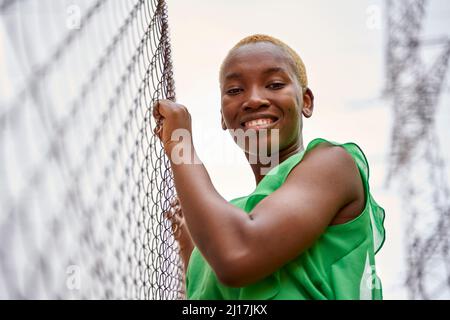 Smiling young woman standing by chainlink fence Stock Photo