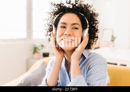 Happy woman with curly hair listening music through wireless headphones at home Stock Photo