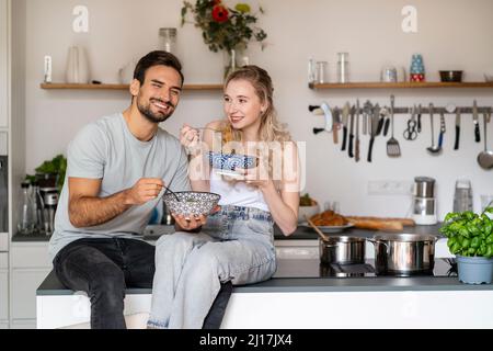 Smiling young woman and man eating spaghetti sitting on kitchen island at home Stock Photo