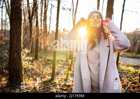 Happy woman in knit hat standing by trees in forest on sunny day Stock Photo