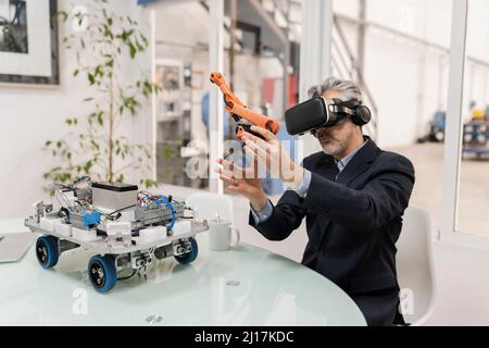 Businessman wearing virtual reality holding robotic arm model sitting at desk Stock Photo