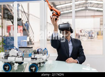 Smiling businessman wearing virtual reality holding robotic arm model sitting at desk Stock Photo