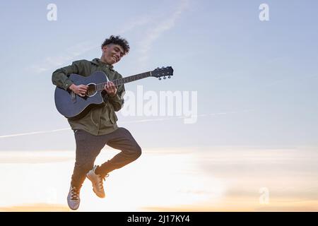 Happy Latin teenager jumping with guitar over the blue sky at sunset. Stock Photo