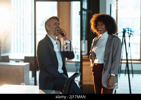 Businessman laughing and talking on mobile phone by colleague at work place Stock Photo