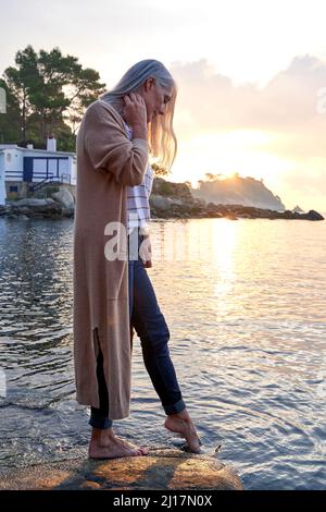 Woman dipping foot in water standing on rock at beach Stock Photo