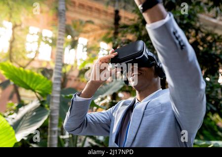 Smiling businessman with hand raised wearing virtual reality simulator in front of plants Stock Photo