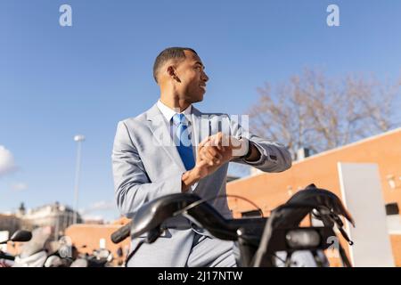Smiling businessman checking time sitting on electric bicycle on sunny day Stock Photo