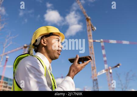 Engineer talking on smart phone through speaker at construction site on sunny day Stock Photo