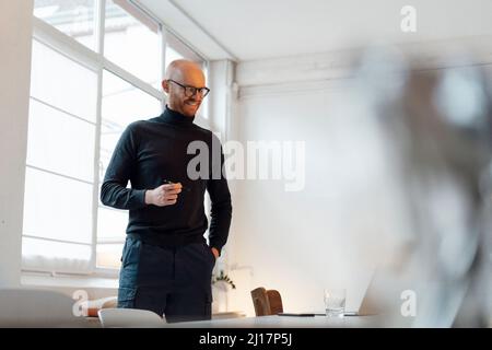 Smiling young businessman standing with hand in pocket at desk Stock Photo
