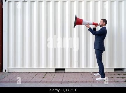 Businessman shouting through megaphone by container Stock Photo