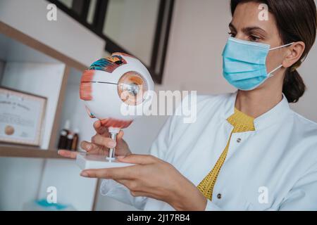 Female doctor with protective face mask holding human eye structure in clinic Stock Photo