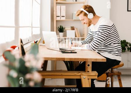 Young man listening music through wireless headphones writing in book siting at table Stock Photo