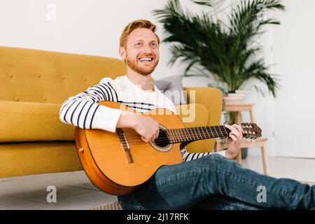 Cheerful man with guitar sitting in front of sofa at home Stock Photo