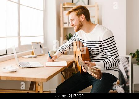 Smiling young man with guitar writing musical notes learning through laptop at home Stock Photo