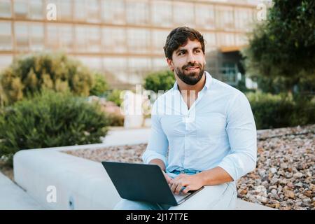 Smiling freelancer with laptop sitting on concrete seat at office park Stock Photo