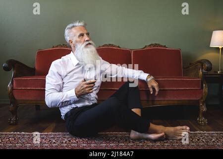 Handsome bearded senior man sitting on the floor and drinking red wine Stock Photo