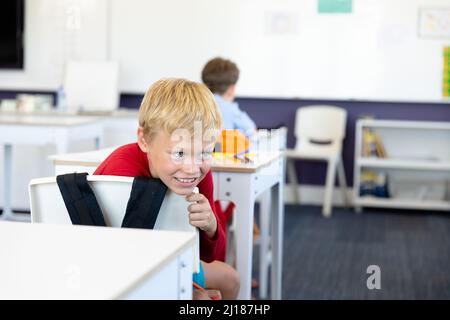 Smiling caucasian elementary schoolboy looking away while leaning on chair in classroom Stock Photo
