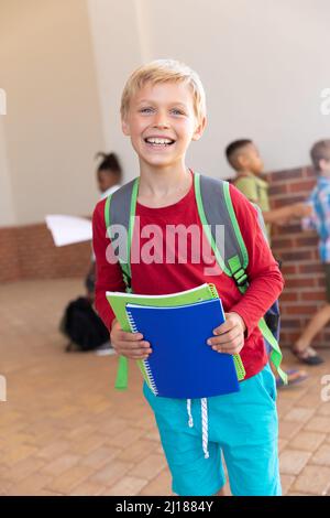 Portrait of smiling caucasian elementary schoolboy with books and backpack standing in school Stock Photo