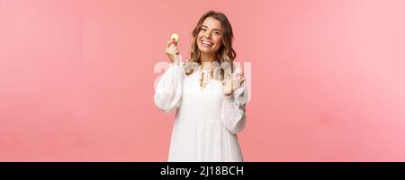 Holidays, spring and party concept. Portrait of tender, lovely blond woman in white dress, dancing joyfully with two macarons, smiling happy eating Stock Photo