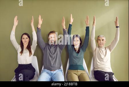 Portrait of people sitting in row on chairs and voting unanimously, raising two hands each. Stock Photo