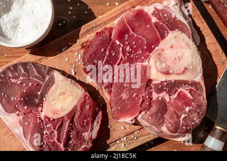 Two steaks of raw beef ossobuco on a wooden board with seasoning to flavour the pieces of meat. Animal protein concept, cheap cuts of beef. Aereal vie Stock Photo