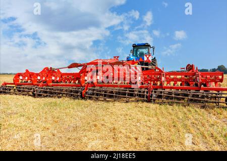 A red multifunctional agricultural harrow stands as a hitch on an agricultural tractor on the stubble of a wheat field on a summer day. ?opy space. Stock Photo