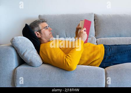 Relaxed middle-aged man with glasses lying on the sofa reading a book. Comfort at home Stock Photo