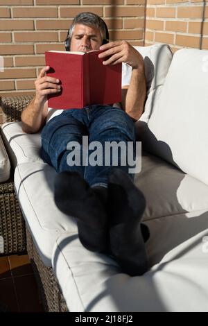 Man lying on a sofa reading a book on a terrace in the sun wearing headphones Stock Photo