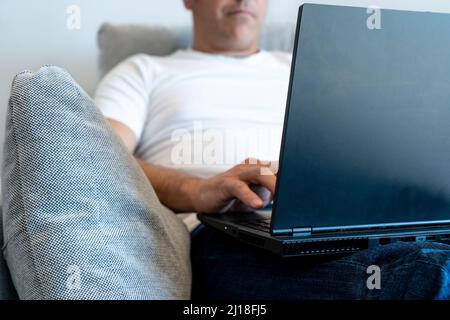 Foreground on unrecognizable man sitting in an armchair using a laptop. Focus on the laptop Stock Photo