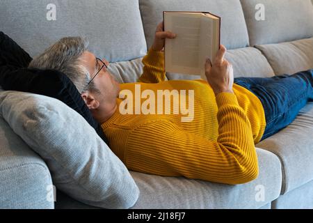 Gray-haired man with glasses lying on the sofa reading a book. Relaxing at home Stock Photo