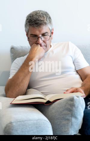 Grey-haired man with glasses sitting in an armchair reading a book. Relax at home concept Stock Photo