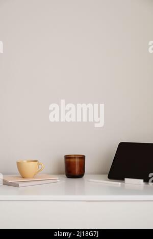 Minimalist home office desk table. Feminine workspace with mug of tea, digital tablet, candle, paper notebooks. White wall background. Nordic, boho st Stock Photo