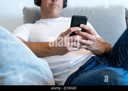 Unrecognizable man on sofa using smartphone and wearing headphones. Hands with phone in foreground Stock Photo