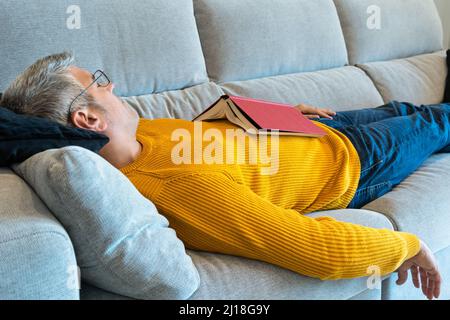 Man with glasses sleeping on the sofa with an open book on his chest. Relaxing at home concept Stock Photo