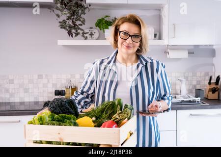 Online home food delivery. Woman checking her online order list on her phone. Wooden box with fresh vegetables and fruits standing on the kitchen tabl Stock Photo