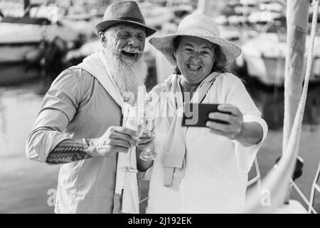 Senior couple doing video call with mobile phone on sailboat during summer vacation - Focus on faces - Black and white edition