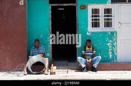 Two men sit while one works making a type of metal tin with wires in Havana, Cuba. Stock Photo