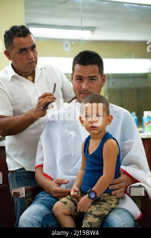 A barber cuts the hair of a father and son at a barbershop in Havana, Cuba. Stock Photo
