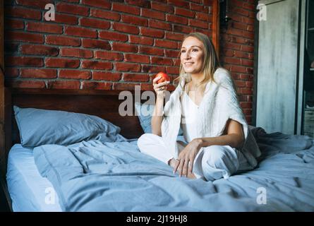 Young adult woman with blonde long hair in casual with apple fruit sitting on bed at home Stock Photo