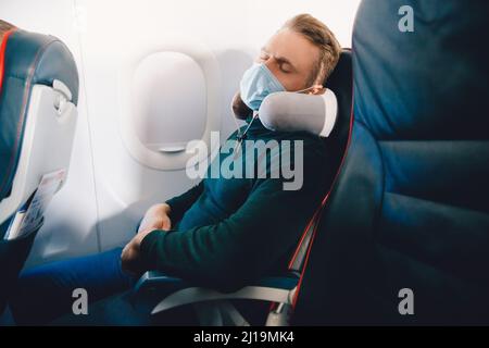 Male passenger in protective mask against viruses of airplane enjoys pillow for sleeping in chair. Concept travel relax plane. Stock Photo