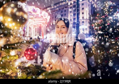 Happy young woman with cup hot coffee walking with bokeh light evening snow. Spending winter vacations in Red square, Moscow, Russia. Stock Photo