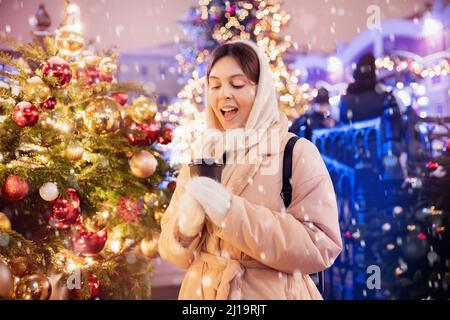 Happy young woman with cup hot coffee walking with bokeh light evening. Spending winter vacations in Red square, Moscow, Russia. Stock Photo