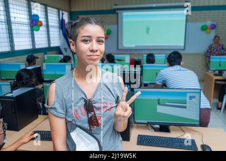 Half-length portrait of a young Latin woman from Central America making a peace symbol with her hand and standing in a classroom full of computers Stock Photo